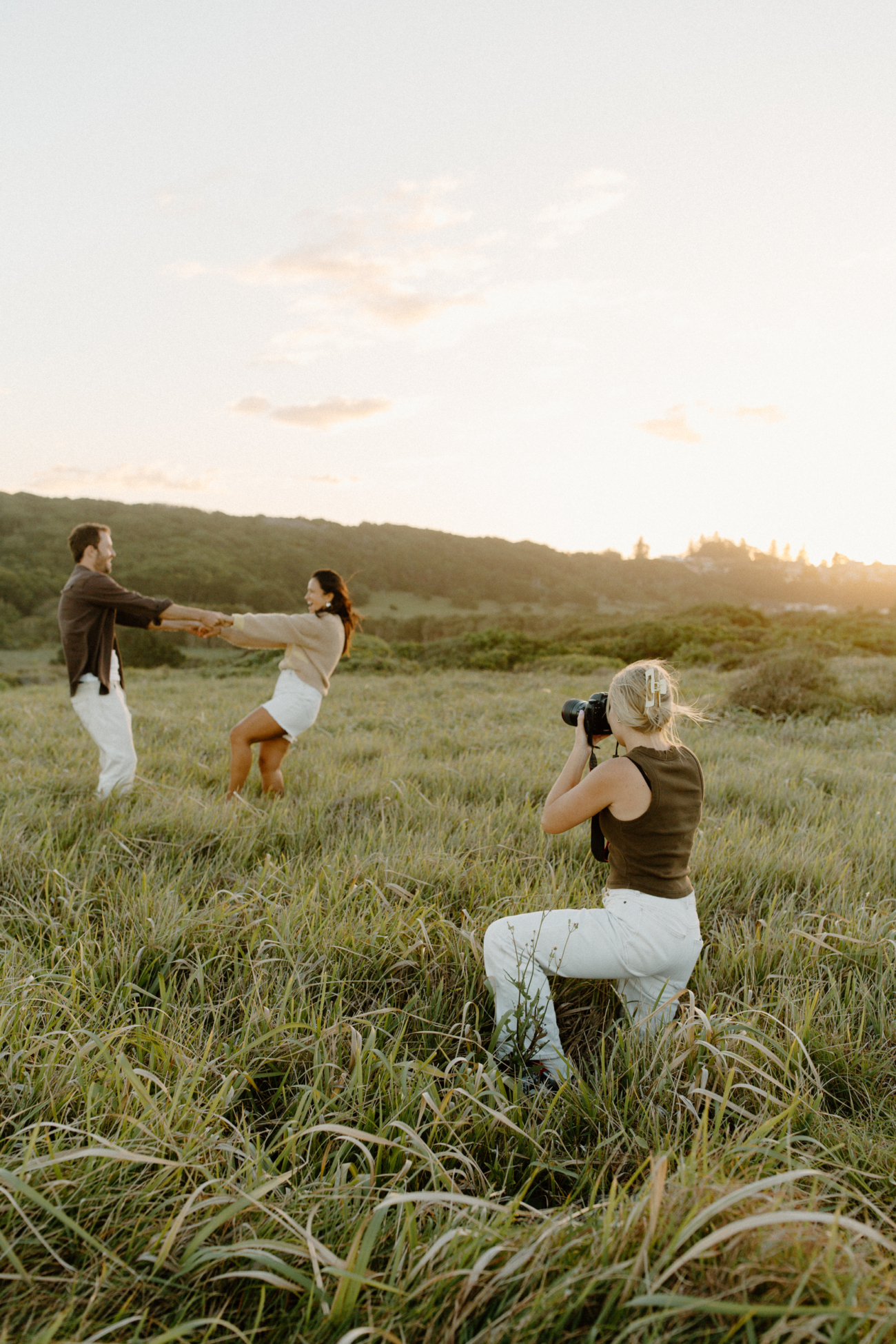 A couple spinning and laughing in long grass while a photographer shoots