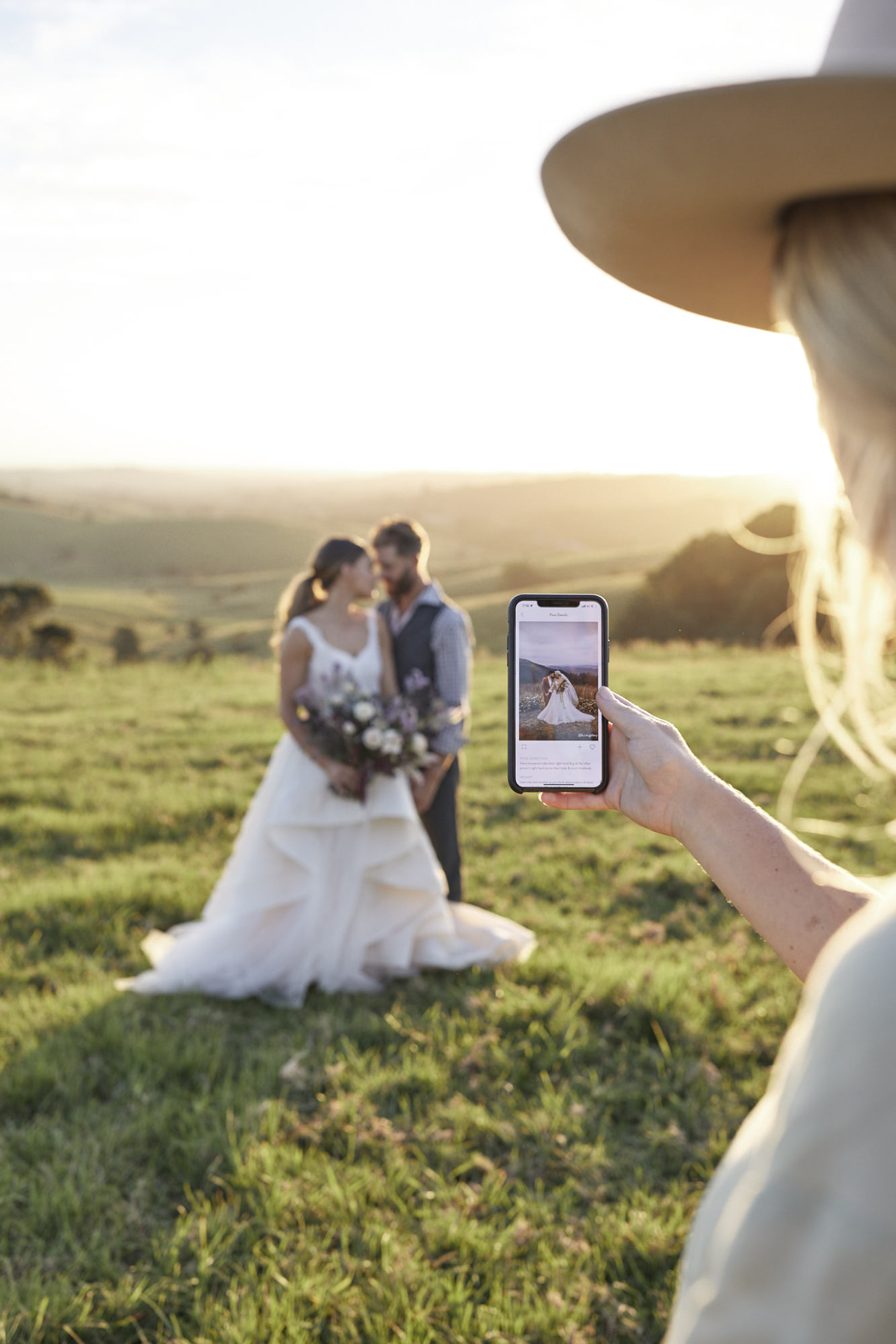 A photographer holding up a phone showing a prompt while a couple stand in the field behind